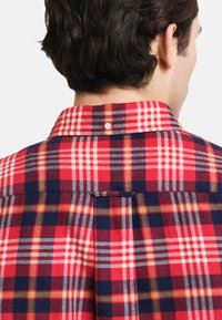 Koszula w czerwono-granatową kratę z kołnierzykiem na guziki, wykonaną z miękkiej bawełny, z dodatkowym plisowaniem z tyłu dla swobody ruchów.