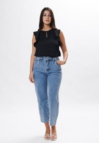 Schwarze ärmellose Bluse mit Rüschenverzierung und Schlüssellochausschnitt, kombiniert mit hellblauen hochgeschnittenen Jeans und weißen Sandalen mit Riemchen.