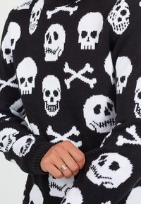 Pull noir avec un motif de crâne et de tibias en blanc. Texture tricotée avec des poignets côtelés et un col ras du cou.