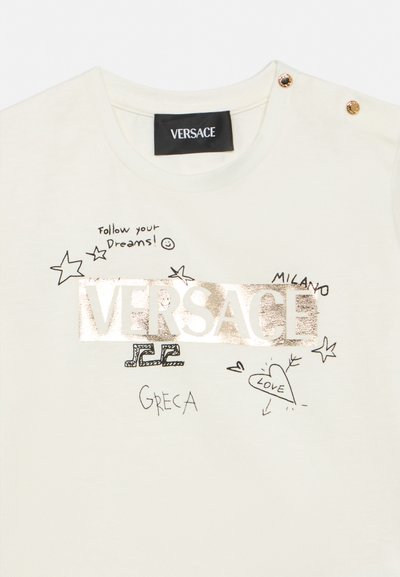 Μπεζ μπλουζάκι με χρυσό λογότυπο VERSACE, μαύρα σχέδια με αστέρια, καρδιά με τη λέξη "LOVE", κείμενο "Ακολούθησε τα Όνειρά σου!" και χρυσά κουμπιά στον ώμο.