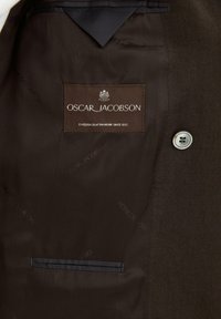 Brązowa podszewka wewnętrzna ubrania z metką Oscar Jacobson z napisem "Szwedzkie rzemiosło od 1903 roku" oraz beżowy guzik z czterema otworami.