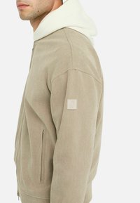 Beige kordfløjl jakke med hvid hætte, lynlås lukning, sidelommer og et lille logo patch på venstre ærme.