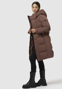 Bruine pufferjas met een hoge kraag en capuchon, voorzien van een rits en een gestikt ontwerp. Gecombineerd met zwarte leggings en veterlaarzen.