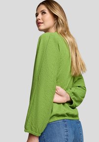 Junge Frau mit langen blonden Haaren, die ein grünes, strukturiertes Langarmoberteil und blaue Jeans trägt. Sie schaut über ihre Schulter vor einem grauen Hintergrund.