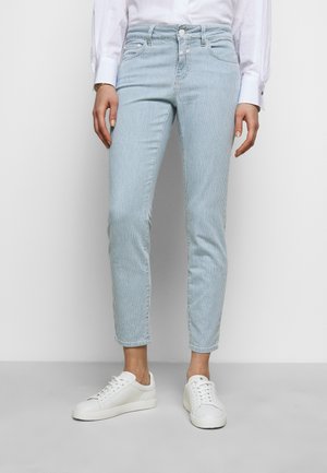 Personne portant un pantalon slim rayé verticalement bleu clair et blanc, une chemise blanche boutonnée et des baskets blanches sur un fond uni.