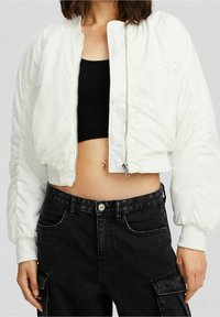 Przycięta biała kurtka puchowa z zapięciem na suwak z przodu, z bufiastymi rękawami i gładką fakturą. Połączona z czarnymi spodniami cargo.