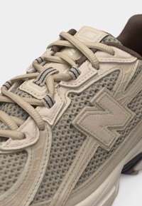 Gros plan d'une basket beige New Balance avec une tige en mesh, des empiècements en suède et des lacets beiges passés dans des œillets en tissu rayé.