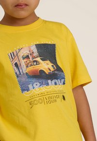 Geel katoenen T-shirt met een afbeelding van een vintage auto op een stenen straat, met tekstoverlay en een levendig ontwerp.