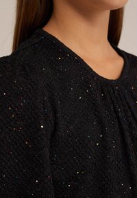 Svart strukturerad tygtröja med glittrande färgglada prickar, med rundad halsringning och draperade detaljer runt axlarna.