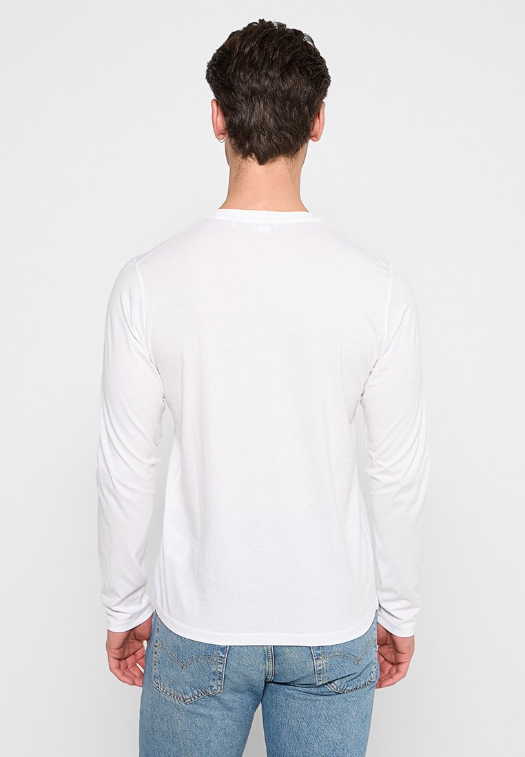 T-shirt bianco a maniche lunghe con collo rotondo. Tessuto morbido, vestibilità slim e orlo che scende sotto la vita. Indossato con jeans azzurri chiari.