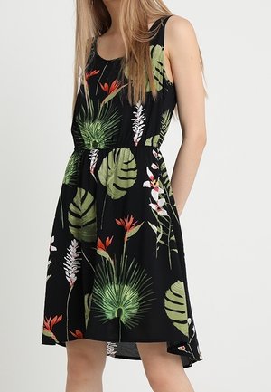 Femme portant une robe noire sans manches avec des imprimés de feuilles et de fleurs tropicales vertes, rouges et blanches, taille cintrée et ourlet arrivant au genou.
