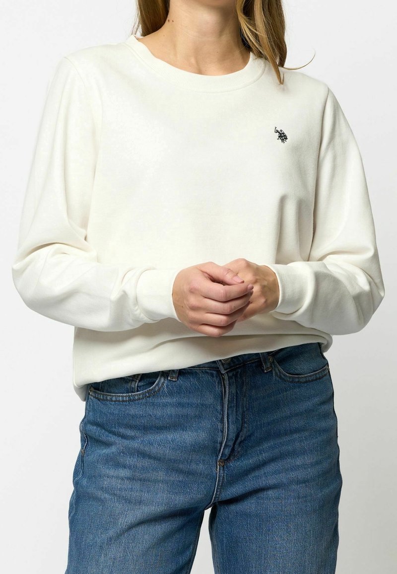 Femme portant un sweat-shirt blanc uni à manches longues avec un petit logo noir brodé sur la poitrine et un jean bleu, mains jointes.