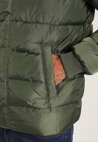 Grön puffjacka med en vadderad design, med en sidficka och ribbade muddar. Tillverkad av ett mjukt, lätt material.