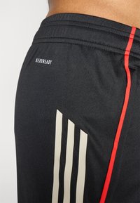Sort atletiske shorts med et struktureret materiale. Bemærkelsesværdige detaljer inkluderer røde sidelister og tre lodrette cremefarvede striber på den nederste del af benet.