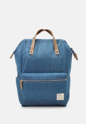 Sac à dos en denim bleu avec des poignées en cuir beige, poche zippée à l'avant et détails cousus. Présente un design structuré et droit.