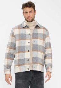 Veste à carreaux gris, crème et marron. Fermeture à boutons sur le devant avec poche poitrine. Texture tissée, col avec col roulé beige assorti.