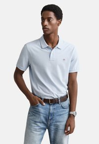 Lichtblauw poloshirt met korte mouwen, voorzien van een kraag en een subtiel logo. Gedragen met lichtblauwe jeans en een bruine riem.