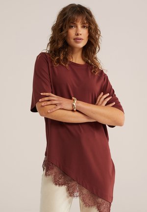 Kobieta z kręconymi włosami, ubrana w asymetryczny bordowy top z koronkową lamówką i złotą bransoletką, stojąca z założonymi ramionami.