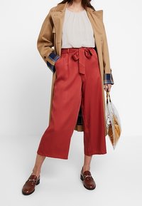 Røde vide bukser med knyttet talje, parret med en beige bluse og en tan trenchcoat, der har blå ternede detaljer på ærmerne.