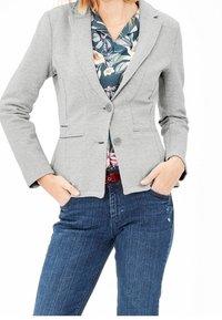 Blazer grigio con finitura testurizzata, caratterizzato da una chiusura a bottone singolo e due tasche frontali, indossato su una camicia floreale e abbinato a jeans blu.
