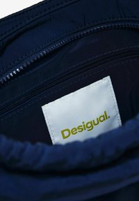 Navyblaue Stoffinneneinrichtung mit zwei Reißverschlusstaschen. Weißes Etikett mit "Desigual." in Grün. Glatte Textur, minimalistisches Design.
