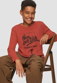 Lächelnder Junge mit kurzen lockigen Haaren, der ein rostfarbenes Langarmshirt trägt, auf dem steht "Schau niemals zurück, es gibt nur 1", sitzt auf einem Holzstuhl.