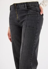 Jeans de mezclilla negra con cremallera, bolsillos laterales y costuras contrastantes. La tela tiene un ligero brillo y un ajuste estructurado.