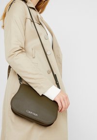 Borsa a tracolla in pelle verde oliva con bordo cucito, caratterizzata da un logo argentato e una tracolla regolabile. Abbinata a un cappotto beige.