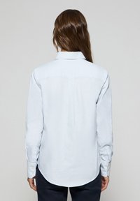 Ljusblå långärmad skjorta med krage, detaljer med veck bak och rundad nederkant. Har knäppte manschetter och en slät textur.