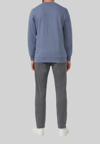 Muž v modrej košeli s dlhým rukávom, šedých džínsoch a bielych teniskách, stojí proti svetlošedému pozadíu.