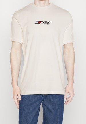 Person wearing a beige Tommy Hilfiger t-shirt with logo on chest and blue textured pants, standing against a plain background.