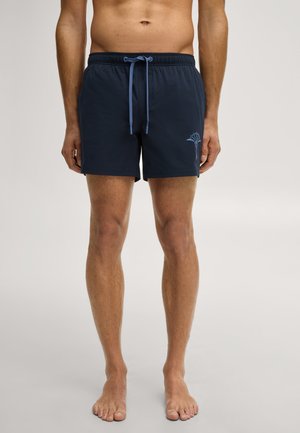 Mann steht barfuß und trägt marineblaue Badeshorts mit hellblauem Kordelzug und Palmenlogo am linken Oberschenkel, einfarbiger heller Hintergrund.