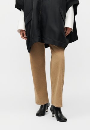 Osoba ubrana w beżowe spodnie o prostych nogawkach, czarne botki na obcasie, białą bluzę z długim rękawem oraz czarną, oversize'ową kurtkę z kieszeniami.