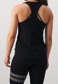 Débardeur de sport noir à dos nageur, fabriqué en tissu doux ; porté avec des leggings noirs dotés de bandes perforées argentées près des mollets.