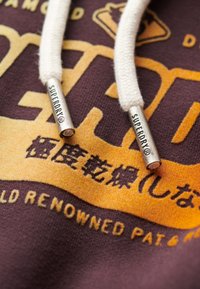 Bluza z kapturem w głębokim kolorze burgundowym, z białymi sznurkami i metalicznymi srebrnymi końcówkami z wytłoczonym napisem "SUPERDRY". Tekst w kolorze żółtym i czarnym.