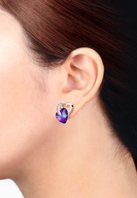 Silverfärgade hjärtformade örhängen med en lila kristall i mitten och små accentstenar, visade på ett öra.