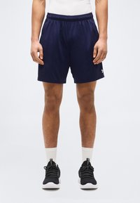 Άνδρας που φοράει ναυτικό μπλε αθλητικό σορτς, λευκές κάλτσες μέχρι τον αστράγαλο και μαύρα παπούτσια τρεξίματος, στέκεται μπροστά σε σκηνικό με απλό ανοιχτό φόντο.