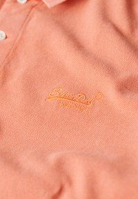 Polo shirt in licht koraal stof, met een gestructureerde weefsel en een opvallend geborduurd logo in geel-oranje op de linkerkant van de borst.