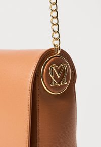 Sac à main en cuir marron avec une chaîne dorée, mettant en avant un accent métallique circulaire proéminent embossé d'un logo "M" stylisé.