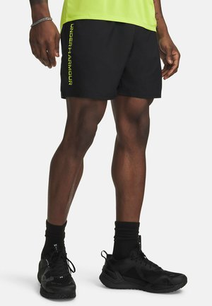 Man staat, draagt zwarte Under Armour-short met geel logo, neon gele top, zwarte hoge sokken, en zwarte sportschoenen op een witte achtergrond.