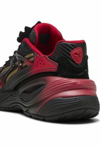 Černé a červené sportovní boty s síťovinou a syntetickými materiály, texturovanou podrážkou a jedinečnými designovými prvky na svršku a patě.