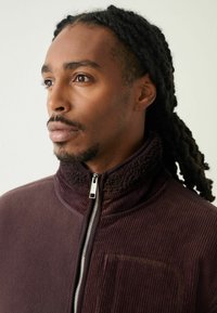 Dark purple zip-up jacket made of soft fleece with ribbed texture and a high collar. Features a silver zipper and a pocket on the chest.