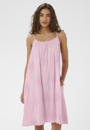 Frau mit lockigem Haar, die ein ärmelloses, locker sitzendes rosa Kleid mit strukturierten kreisförmigen Mustern trägt und vor einem einfarbigen Hintergrund steht.