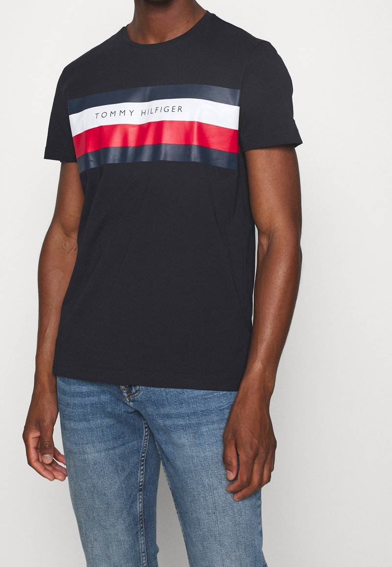 Černé bavlněné tričko s horizontálním pruhovaným designem v modré, červené a bílé barvě, s nápisem "TOMMY HILFIGER" vytištěným uprostřed.