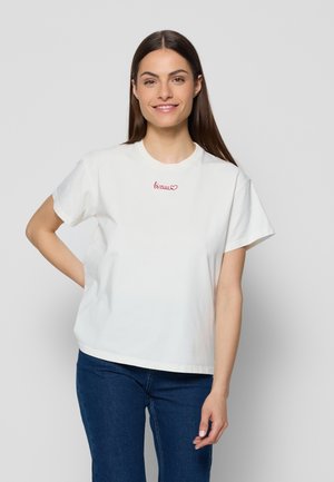 Jonge vrouw met lang bruin haar, die een wit T-shirt met rode tekst en donkerblauwe spijkerbroek draagt, staand tegen een effen lichte achtergrond.