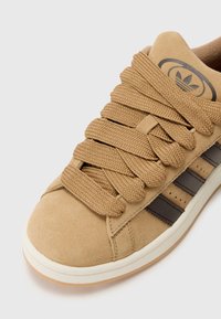 Tan suede sneaker with textured, thick laces and a white rubber sole. Features three dark brown stripes and an embossed logo on the tongue.
