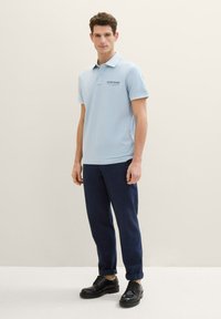 Svetlo modra polo majica s kratkimi rokavi, ovratnikom in detajlom logotipa; v kombinaciji z mornarsko modrimi hlačami in črnimi vezalnimi čevlji. Gladka tekstura.