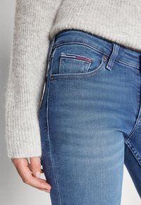 Jeans de mezclilla azul de tiro alto con una textura suave. Cuentan con un pequeño bolsillo frontal con costuras decorativas en rojo, blanco y azul.