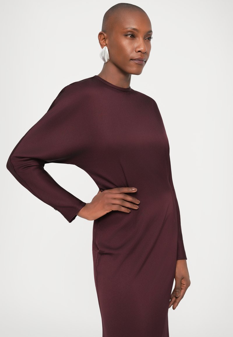 Bordeauxfarbenes Langarmkleid mit hohem Halsauschnitt, glattem Stoff und lockerer Silhouette, kombiniert mit strukturierten silbernen Ohrringen.