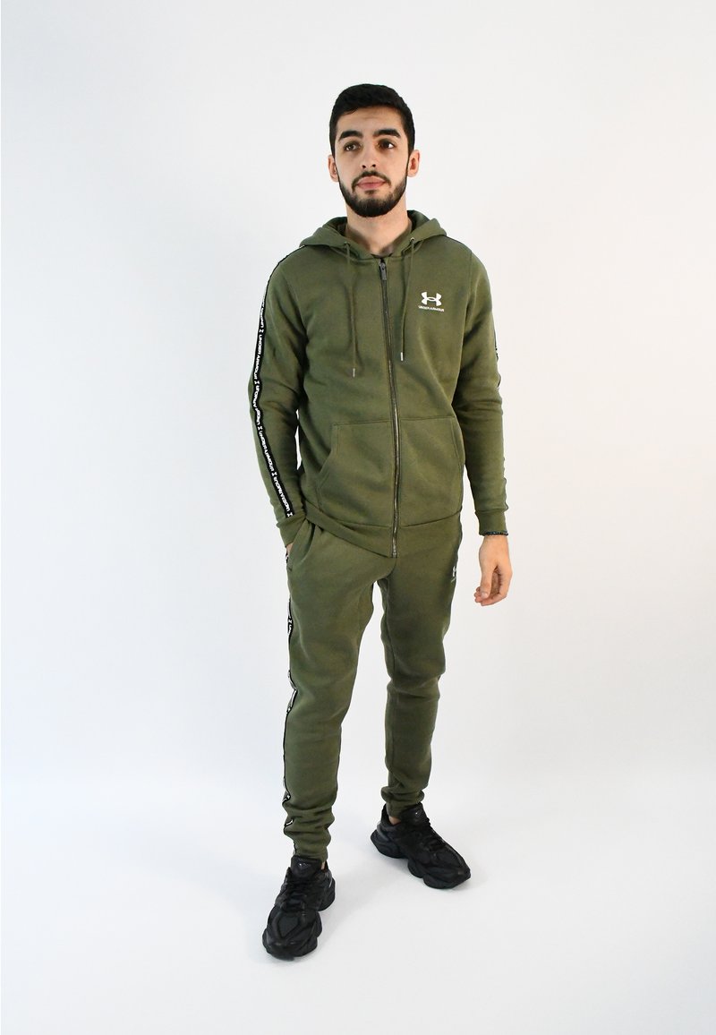 Conjunto de sudadera con capucha y pantalones jogger en verde oliva, con cierre de cremallera frontal, rayas laterales y dos bolsillos delanteros. Combinado con zapatillas negras sobre un fondo liso.